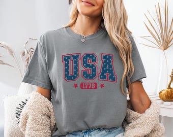 May include: A grey short-sleeved t-shirt featuring the text USA in large blue block letters with a white star pattern, outlined in red. Below the text, the number 1776 is printed in red, flanked by two red stars on a grey background.