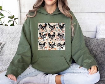 Op de afbeelding: Een olijfgroene hoodie met een centrale grafische print bestaande uit negen vierkante panelen. Elk paneel toont een zwart silhouet van een kip gevuld met een bloemenpatroon, tegen een achtergrond van gedempte oranje en blauwe bloemen.
