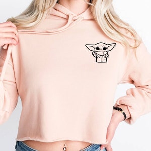 May include: A light pink cropped hoodie with a black outline of a character from the Star Wars franchise, "The Mandalorian", on the front left side of the hoodie.