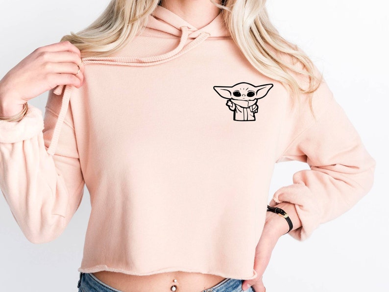 May include: A light pink cropped hoodie with a black outline of a character from the Star Wars franchise, "The Mandalorian", on the front left side of the hoodie.