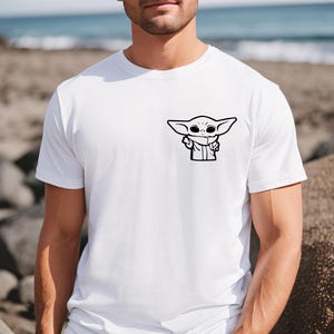 May include: A white t-shirt with a black and white cartoon image of a character from the Star Wars franchise, The Mandalorian, known as "Baby Yoda".