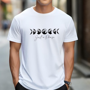 May include: White t-shirt featuring a black graphic of moon phases with the text "Just a Phase" in a script font. The shirt is a crew neck and short-sleeved.