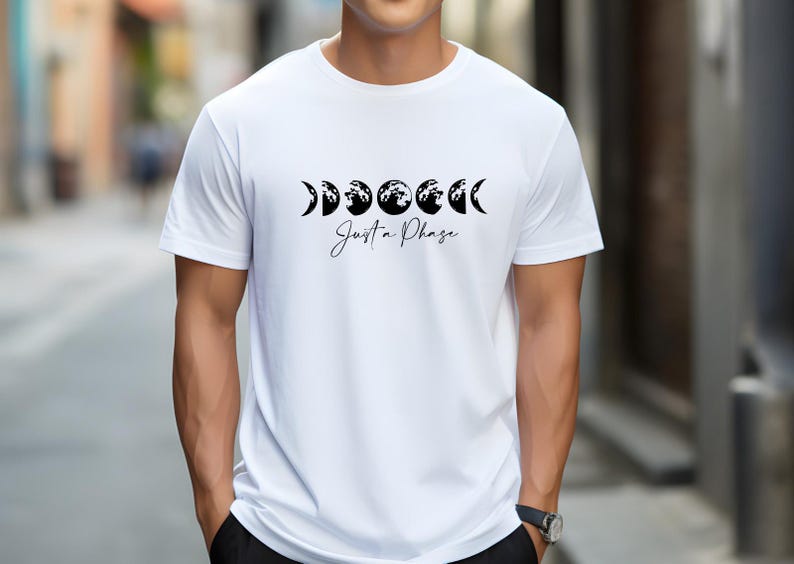 May include: White t-shirt featuring a black graphic of moon phases with the text "Just a Phase" in a script font. The shirt is a crew neck and short-sleeved.