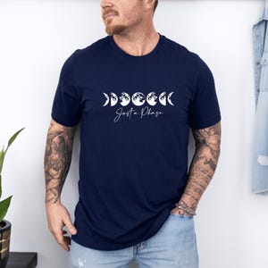 May include: Navy blue t-shirt featuring a white graphic of the phases of the moon and the text "Just a Phase". The design is centered on the front of the shirt. The shirt has a crew neck and short sleeves.