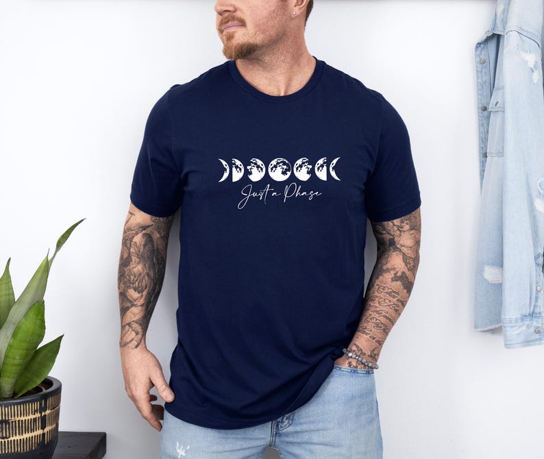 May include: Navy blue t-shirt featuring a white graphic of the phases of the moon and the text "Just a Phase". The design is centered on the front of the shirt. The shirt has a crew neck and short sleeves.