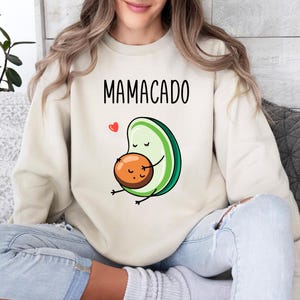 May include: A cream-coloured crewneck sweatshirt featuring a graphic of a stylised avocado with a face, holding a round pit, with the text MAMACADO printed in black above the illustration and a small red heart icon to the left of the avocado.