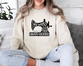 May include: A cream-colored crewneck sweatshirt featuring a black graphic of a sewing machine. The text reads: She works with her hands in delight, Proverbs 31:13. The design is centered on the chest of the long-sleeved pullover garment.