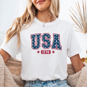 May include: A white short-sleeved t-shirt featuring the text USA in large, distressed blue block letters filled with white stars. Below the text, the number 1776 is printed in a red rectangular box, flanked by two small red stars.