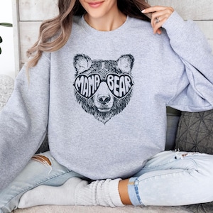 May include: A light grey crewneck sweatshirt featuring a black graphic print of a bear wearing heart-shaped sunglasses. The text "MAMA BEAR" is integrated into the lenses of the sunglasses. The sweatshirt has long sleeves and a ribbed neckline.