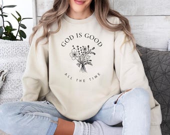 May include: A cream-colored crewneck sweatshirt featuring a black line-art illustration of a wildflower bouquet in the center. The text "GOD IS GOOD" is printed in a curved arch above the flowers, and "ALL THE TIME" is printed below the bouquet.