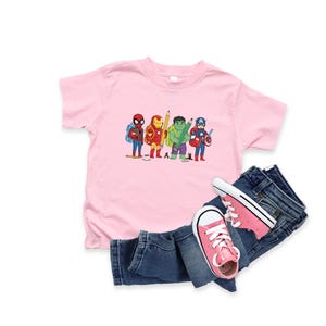 May include: A pink t-shirt featuring a cartoon illustration of Spider-Man, Iron Man, Hulk, and Captain America with pencils. The shirt is paired with blue denim jeans and pink sneakers.
