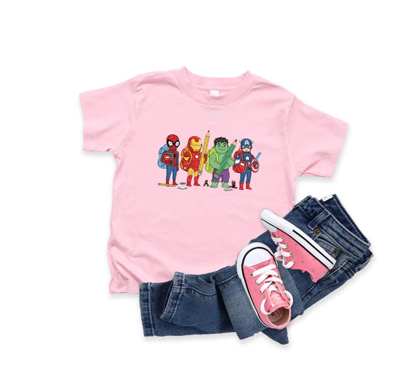 May include: A pink t-shirt featuring a cartoon illustration of Spider-Man, Iron Man, Hulk, and Captain America with pencils. The shirt is paired with blue denim jeans and pink sneakers.