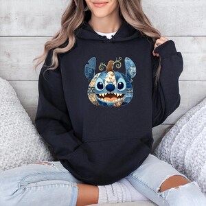 K&ouml;nnte beinhalten: Ein marineblauer Kapuzenpullover mit einem von Stitch inspirierten K&uuml;rbisdesign. Der K&uuml;rbis hat blaue, braune und wei&szlig;e Patchwork-Details mit Stitchs Gesicht und Ohren. Der Hoodie wird mit hellblauen Jeans getragen.