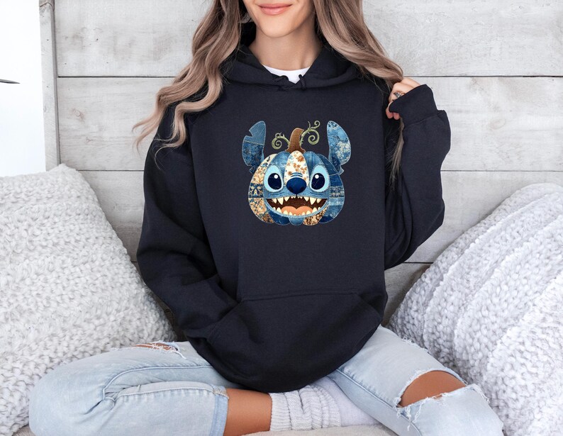 K&ouml;nnte beinhalten: Ein marineblauer Kapuzenpullover mit einem von Stitch inspirierten K&uuml;rbisdesign. Der K&uuml;rbis hat blaue, braune und wei&szlig;e Patchwork-Details mit Stitchs Gesicht und Ohren. Der Hoodie wird mit hellblauen Jeans getragen.