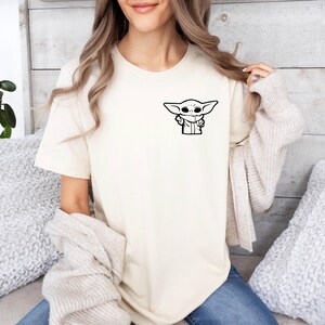 May include: A white t-shirt with a black and white illustration of a character from the Star Wars franchise, The Mandalorian, known as "The Child" or "Baby Yoda".