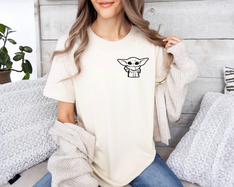 May include: A white t-shirt with a black and white illustration of a character from the Star Wars franchise, The Mandalorian, known as "The Child" or "Baby Yoda".