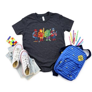May include: A charcoal gray t-shirt featuring a colorful graphic of superheroes with pencils. The image includes a blue backpack, white sneakers, a Rubik's cube, and colorful pencils, suggesting a back-to-school theme.