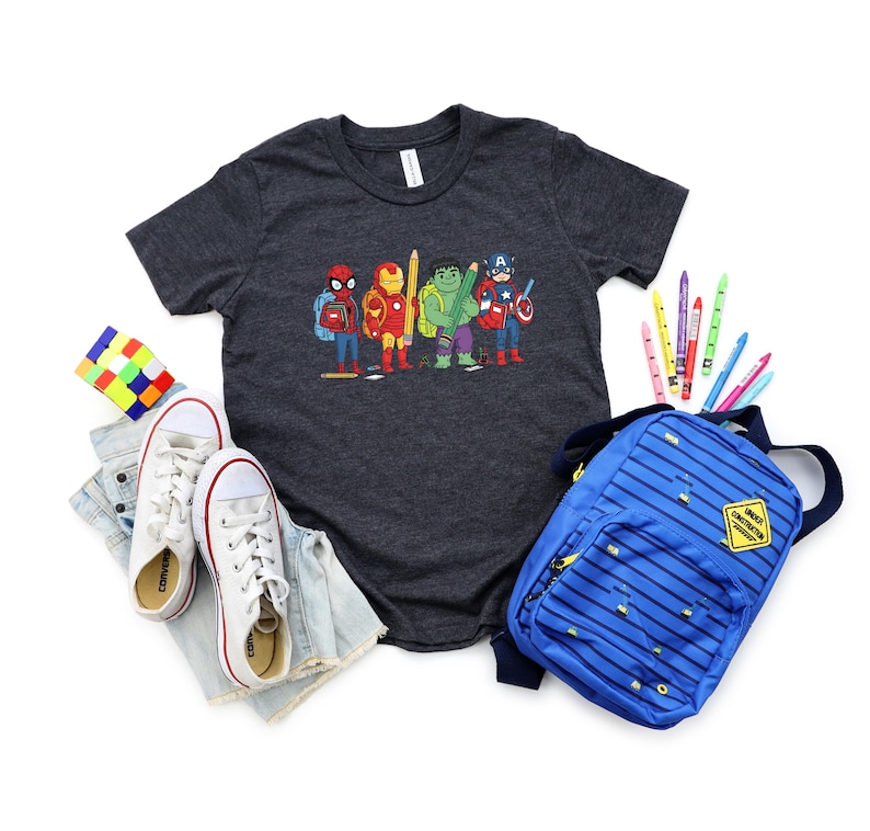 May include: A charcoal gray t-shirt featuring a colorful graphic of superheroes with pencils. The image includes a blue backpack, white sneakers, a Rubik's cube, and colorful pencils, suggesting a back-to-school theme.