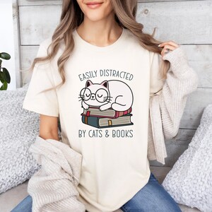 May include: A white t-shirt with a graphic of a cat wearing glasses and sleeping on a stack of books. The text on the shirt reads "Easily Distracted by Cats & Books".