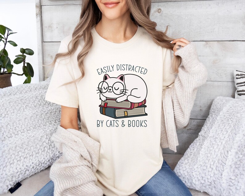 May include: A white t-shirt with a graphic of a cat wearing glasses and sleeping on a stack of books. The text on the shirt reads "Easily Distracted by Cats & Books".