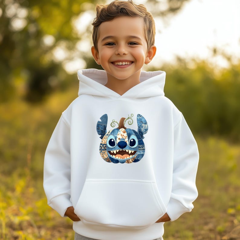 K&ouml;nnte beinhalten: Ein wei&szlig;er Kapuzenpullover mit einem blauen und beigefarbenen Stitch-K&uuml;rbis-Design. Die Grafik hat ein Patchwork-Design und ein l&auml;chelndes Gesicht. Der Pullover hat eine Vordertasche und eine Kapuze mit Kordelzug.