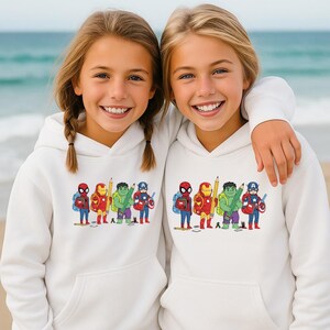 May include: Two young girls wearing white hoodies with a colorful graphic of superheroes as students. The graphic includes Spider-Man, Iron Man, Hulk, and Captain America with backpacks and pencils. The girls are smiling on a beach.