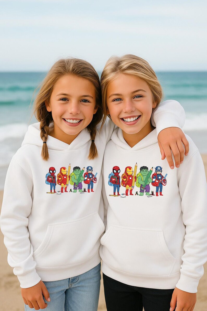 May include: Two young girls wearing white hoodies with a colorful graphic of superheroes as students. The graphic includes Spider-Man, Iron Man, Hulk, and Captain America with backpacks and pencils. The girls are smiling on a beach.