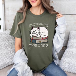May include: A green t-shirt with a white cat wearing glasses and sleeping on a stack of books. The text on the shirt reads "Easily Distracted by Cats & Books".