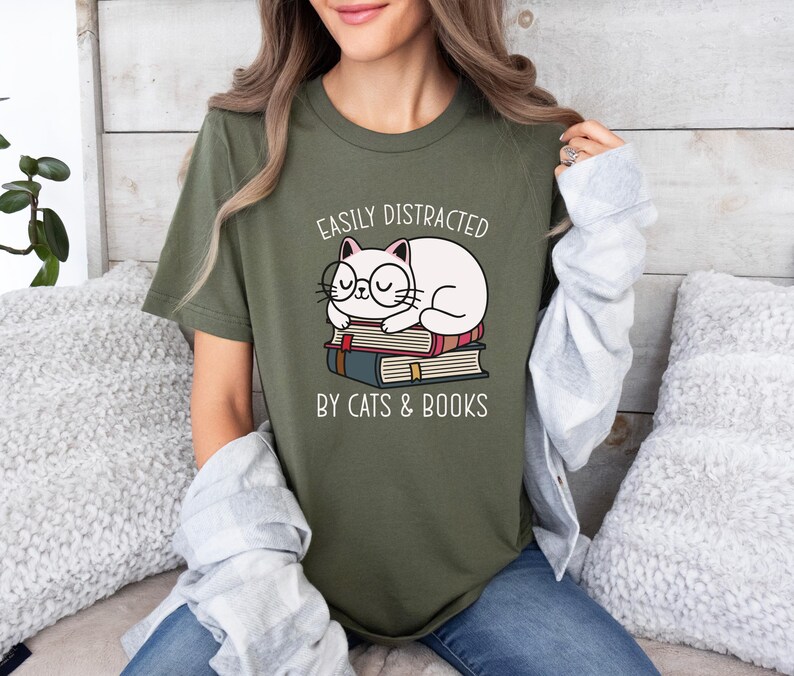 May include: A green t-shirt with a white cat wearing glasses and sleeping on a stack of books. The text on the shirt reads "Easily Distracted by Cats & Books".