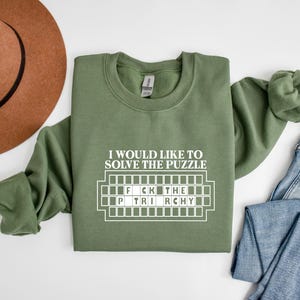 Puede incluir: Una sudadera de cuello redondo color verde oliva con texto blanco que dice I WOULD LIKE TO SOLVE THE PUZZLE sobre un gráfico de un tablero de juego con las letras F C K THE P TRI RCHY rellenas, creando un diseño tipo rompecabezas.