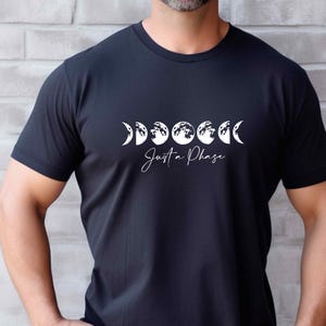 May include: Black t-shirt featuring a white graphic of moon phases. The text "Just a Phase" is written in a cursive font below the moon phases. The shirt is short-sleeved and appears to be made of a soft material.
