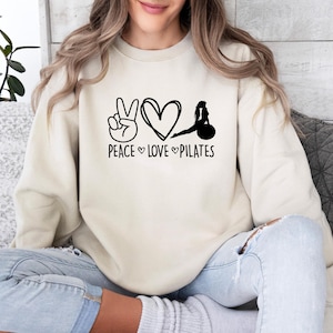 May include: A beige crewneck sweatshirt featuring a black graphic print on the chest. The design includes a peace sign, a heart, and a silhouette of a person on a pilates ball, with the text "PEACE LOVE PILATES" printed in black below.