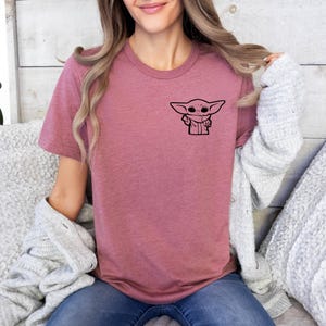 May include: A mauve t-shirt with a black graphic of a character from the Star Wars franchise. The character is known as "The Child" or "Baby Yoda".