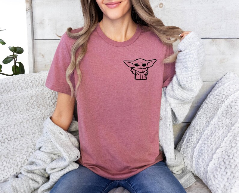 May include: A mauve t-shirt with a black graphic of a character from the Star Wars franchise. The character is known as "The Child" or "Baby Yoda".