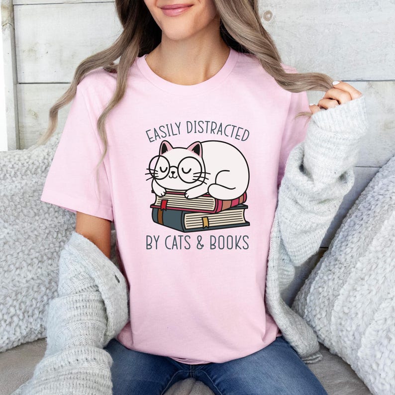May include: Pink t-shirt with a white cat wearing glasses and sleeping on a stack of books. The text on the shirt reads "Easily Distracted by Cats & Books."