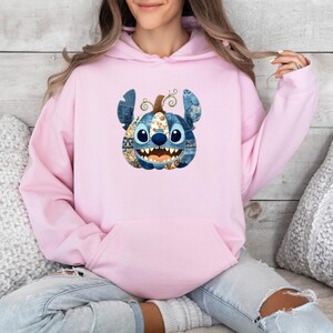 K&ouml;nnte beinhalten: Ein hellrosa Kapuzenpullover mit einer Grafik von Stitch als K&uuml;rbis. Der Stitch-K&uuml;rbis hat blaue Jeans-Ohren, einen braunen Stiel und ein l&auml;chelndes Gesicht mit scharfen Z&auml;hnen. Der Hoodie hat eine K&auml;ngurutasche.