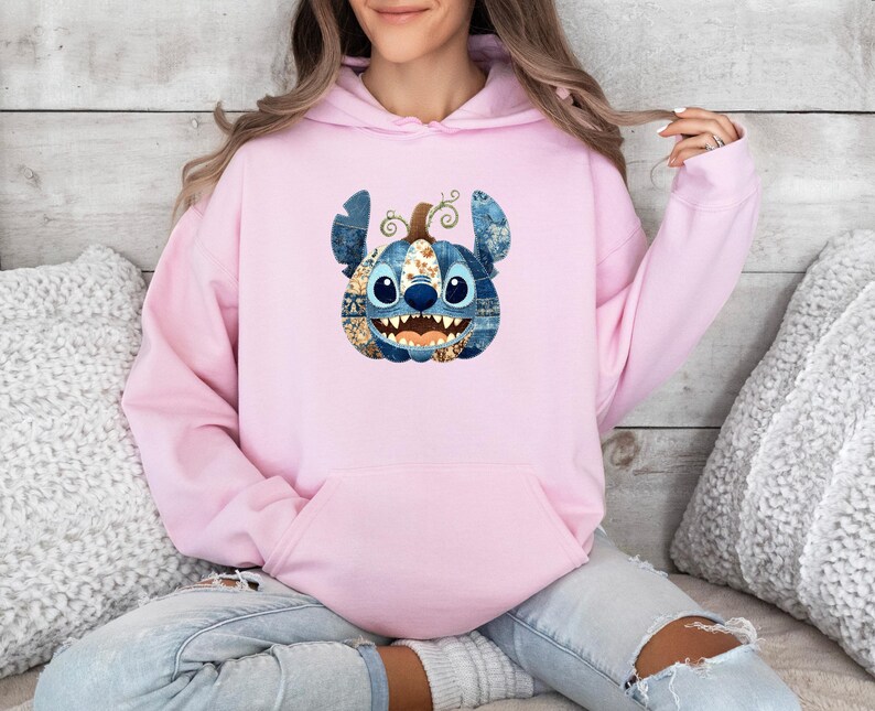 K&ouml;nnte beinhalten: Ein hellrosa Kapuzenpullover mit einer Grafik von Stitch als K&uuml;rbis. Der Stitch-K&uuml;rbis hat blaue Jeans-Ohren, einen braunen Stiel und ein l&auml;chelndes Gesicht mit scharfen Z&auml;hnen. Der Hoodie hat eine K&auml;ngurutasche.