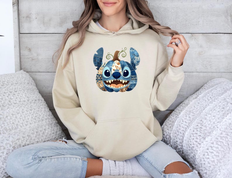 K&ouml;nnte beinhalten: Beiger Kapuzenpullover mit einem Stitch-K&uuml;rbis-Motiv. Das K&uuml;rbismotiv hat blaue, jeansartige Ohren und ein Patchwork-Gesicht mit breitem Grinsen. Der Hoodie hat eine K&auml;ngurutasche und eine Kapuze mit Kordelzug.