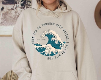May include: A beige hooded sweatshirt featuring a circular graphic on the chest. The design depicts a stylized blue ocean wave with white foam and a mountain peak, surrounded by the text: When you go through deep waters, I will be with you.