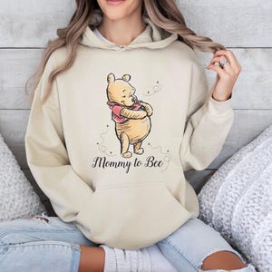 Puede incluir: Una sudadera con capucha de color beige que presenta una ilustración gráfica de Winnie the Pooh con una camiseta roja. Debajo del personaje, el texto Mommy to Bee está impreso en cursiva negra, junto a pequeñas líneas discontinuas que representan abejas.