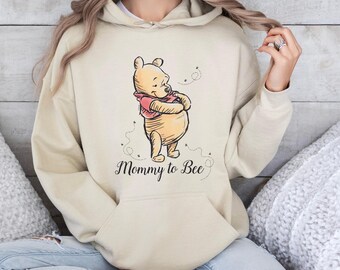 May include: A beige hooded sweatshirt featuring a graphic illustration of Winnie the Pooh wearing a red shirt. Below the character, the text Mommy to Bee is printed in a black cursive script, accompanied by small dashed lines representing flying bees.
