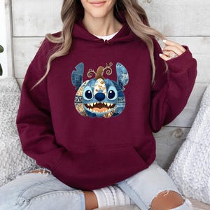K&ouml;nnte beinhalten: Burgunderfarbener Kapuzenpullover mit einem von Stitch inspirierten K&uuml;rbisdesign. Der K&uuml;rbis hat blaue und orangefarbene Patchwork-Details, ein l&auml;chelndes Gesicht und einen lockigen Stiel. Der Hoodie hat eine Kapuze mit Kordelzug und eine K&auml;ngurutasche.
