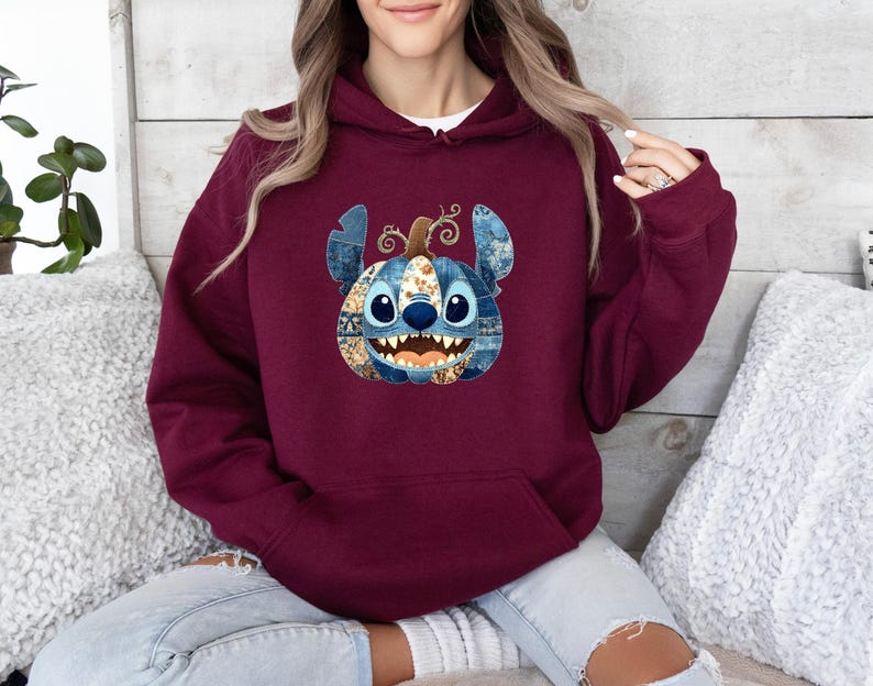 K&ouml;nnte beinhalten: Burgunderfarbener Kapuzenpullover mit einem von Stitch inspirierten K&uuml;rbisdesign. Der K&uuml;rbis hat blaue und orangefarbene Patchwork-Details, ein l&auml;chelndes Gesicht und einen lockigen Stiel. Der Hoodie hat eine Kapuze mit Kordelzug und eine K&auml;ngurutasche.