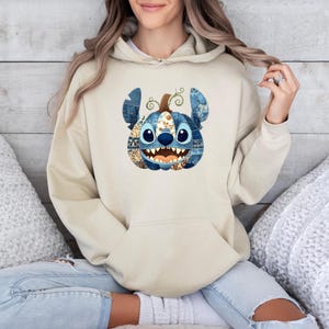 May include: A beige hooded sweatshirt featuring a central graphic of a blue character shaped like a pumpkin. The character has large ears, a wide grin with visible teeth, and a patchwork pattern of denim and floral prints in shades of blue and orange.