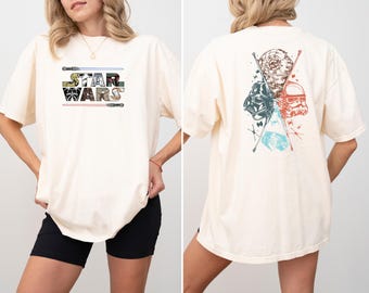 May include: Cream-colored t-shirt featuring the text "STAR WARS" in a colorful, stylized font with lightsabers above and below. The back of the shirt displays a graphic of iconic characters and symbols from the Star Wars saga.