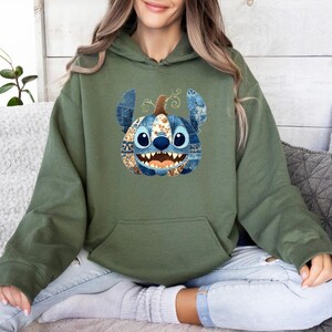 K&ouml;nnte beinhalten: Olivgr&uuml;ner Kapuzenpullover mit einem Stitch-inspirierten K&uuml;rbisdesign. Der K&uuml;rbis hat blaue Jeans- und gemusterte Patches, mit gro&szlig;en blauen Augen und einem grinsenden Mund. Der Hoodie hat eine K&auml;ngurutasche und eine Kapuze mit Kordelzug.