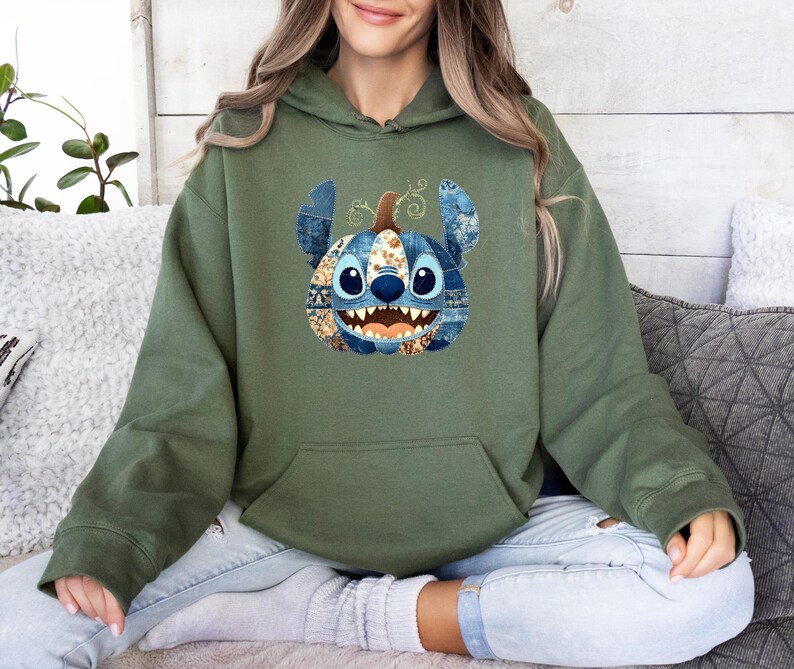 K&ouml;nnte beinhalten: Olivgr&uuml;ner Kapuzenpullover mit einem Stitch-inspirierten K&uuml;rbisdesign. Der K&uuml;rbis hat blaue Jeans- und gemusterte Patches, mit gro&szlig;en blauen Augen und einem grinsenden Mund. Der Hoodie hat eine K&auml;ngurutasche und eine Kapuze mit Kordelzug.