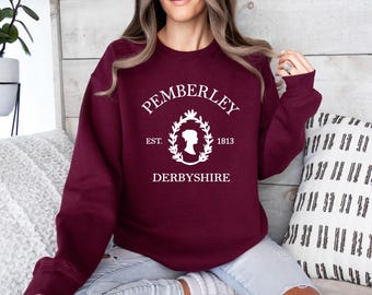 Op de afbeelding: Een bordeauxrode sweater met ronde hals en een witte grafische print op de borst. Het ontwerp bevat de tekst PEMBERLEY, EST. 1813 en DERBYSHIRE, rondom een centraal silhouet van een persoon omlijst door een decoratieve lauwerkrans.