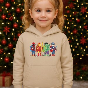 May include: A tan hoodie featuring a colorful embroidered design of cartoon superheroes. The design includes Spider-Man, Iron Man, a pencil, the Hulk, and Captain America. The hoodie has a front pocket and a hood. The background includes a Christmas tree.