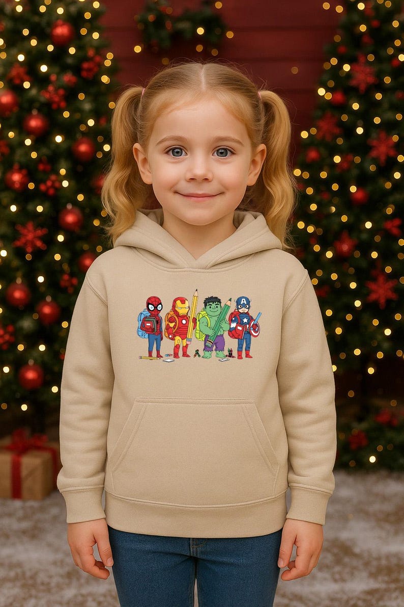 May include: A tan hoodie featuring a colorful embroidered design of cartoon superheroes. The design includes Spider-Man, Iron Man, a pencil, the Hulk, and Captain America. The hoodie has a front pocket and a hood. The background includes a Christmas tree.